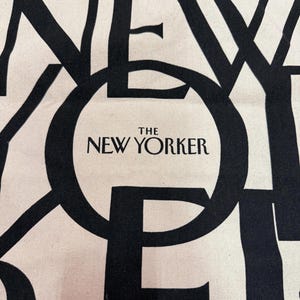 May include: Off-white tote bag with large black text that reads "NEW YORKER" in a bold, overlapping font. The words "THE NEW YORKER" are centered within a black circle. The bag has a simple, minimalist design.