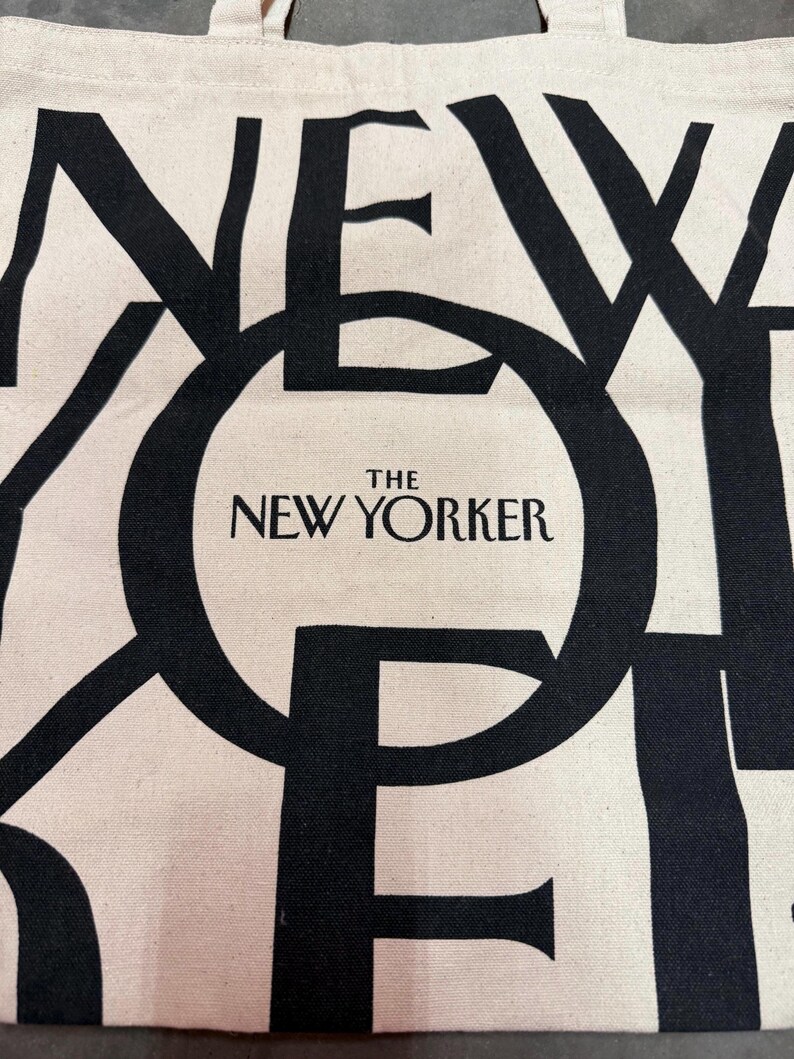 May include: Off-white tote bag with large black text that reads "NEW YORKER" in a bold, overlapping font. The words "THE NEW YORKER" are centered within a black circle. The bag has a simple, minimalist design.