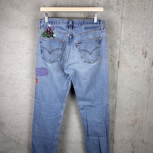 May include: Light blue denim jeans featuring a classic five-pocket design with a high waist and straight legs. The back of the jeans includes a patchwork detail on the left pocket and a small purple patterned fabric patch on the upper left leg.