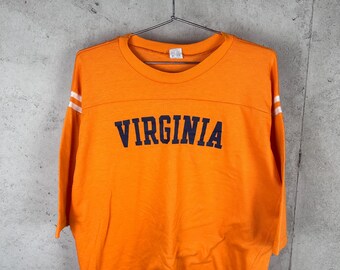 May include: A bright orange short-sleeve t-shirt featuring the word VIRGINIA printed in dark blue block letters across the chest. The sleeves have two horizontal white stripes near the shoulders, and the shirt is displayed on a metal hanger.