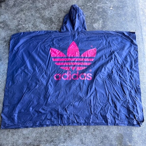 May include: A navy blue Adidas poncho with a hood. The poncho features a large pink Adidas logo and the Adidas wordmark in pink. The material appears to be a lightweight, water-resistant fabric, suitable for outdoor wear.