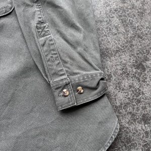 May include: Close-up of a gray button-down shirt. The shirt features a buttoned cuff with two brown buttons. The fabric has a slightly worn appearance, with visible seams and stitching details. The shirt is made of a durable material.