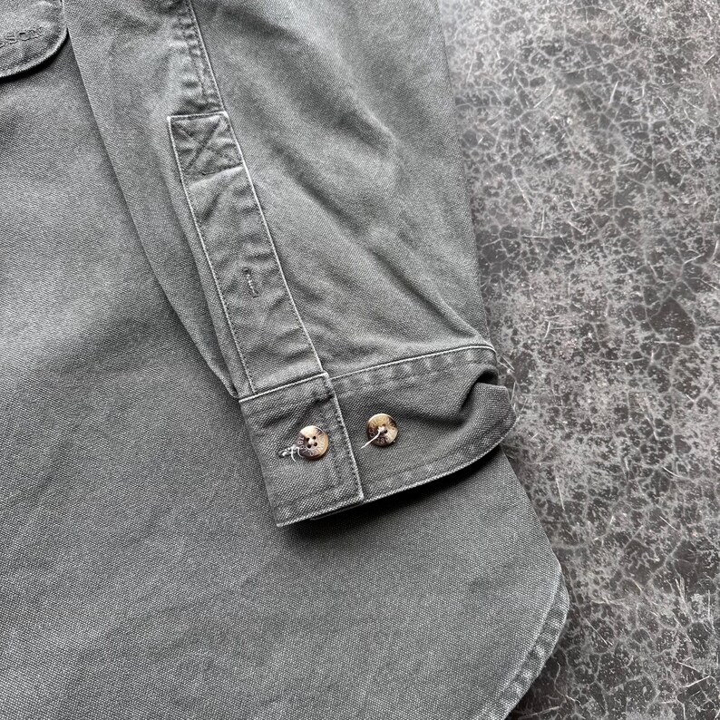 May include: Close-up of a gray button-down shirt. The shirt features a buttoned cuff with two brown buttons. The fabric has a slightly worn appearance, with visible seams and stitching details. The shirt is made of a durable material.