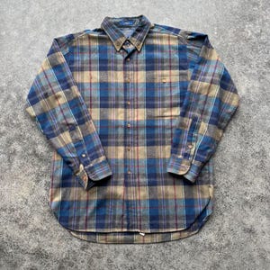 May include: A long-sleeved, button-down flannel shirt in a plaid pattern. The shirt features a color palette of blue, tan, and burgundy. It has a button-down collar, a chest pocket, and buttoned cuffs. The shirt is laid flat on a textured surface.