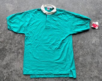 May include: A teal rugby shirt with a white collar and a small logo on the collar. The shirt has short sleeves and a three-button placket. A small tag is attached to the shirt.