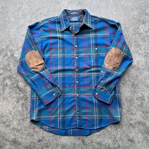 Op de afbeelding: Een blauw, rood en groen geruit flanellen overhemd met bruine elleboogstukken. Het shirt met lange mouwen heeft een knoopsluiting, een borstzak en een kraag. Het shirt is gemaakt van een zacht, comfortabel materiaal.