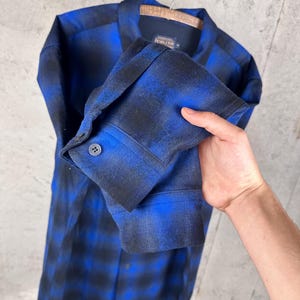 May include: A long-sleeve button-down shirt featuring a blue and black ombre plaid pattern. The shirt has a classic collar, a single buttoned cuff on the sleeve, and a Pendleton brand label visible on the inside of the collar neckline.