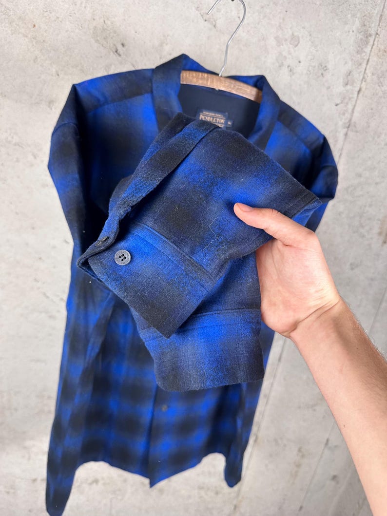 May include: A long-sleeve button-down shirt featuring a blue and black ombre plaid pattern. The shirt has a classic collar, a single buttoned cuff on the sleeve, and a Pendleton brand label visible on the inside of the collar neckline.