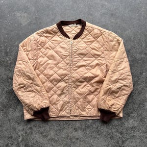 May include: A tan quilted bomber jacket with a brown collar and cuffs. The jacket has a diamond pattern and a full-length zipper. The jacket is a vintage style.