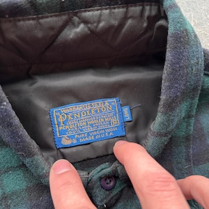 May include: A green and navy plaid shirt with a black quilted lining. The shirt features a blue label with the Pendleton logo and text indicating it's made of pure virgin wool in the USA. The shirt has a button closure.