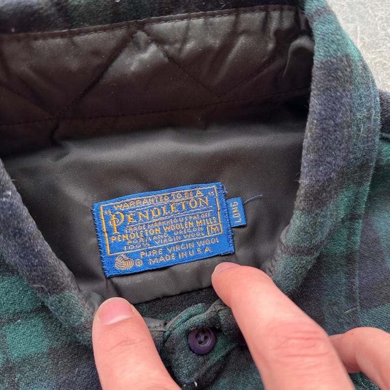 May include: A green and navy plaid shirt with a black quilted lining. The shirt features a blue label with the Pendleton logo and text indicating it's made of pure virgin wool in the USA. The shirt has a button closure.