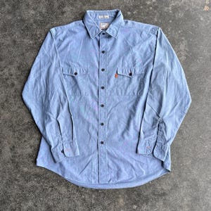 May include: A light blue button-down shirt with two chest pockets and long sleeves. The shirt has black buttons and orange stitching on the cuffs and a small orange tag on the pocket. The shirt is made of a soft, textured fabric.