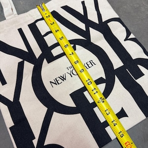 May include: A beige tote bag with black text that reads "The New Yorker." The bag is measured with a yellow tape measure, showing a length of approximately 16 inches. The bag has two beige handles.