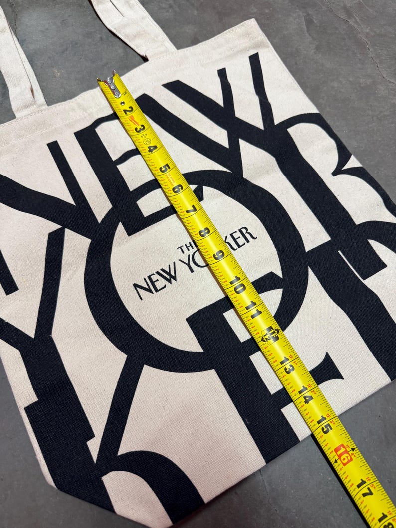 May include: A beige tote bag with black text that reads "The New Yorker." The bag is measured with a yellow tape measure, showing a length of approximately 16 inches. The bag has two beige handles.