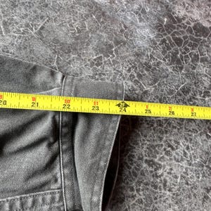 May include: Close-up of a dark gray denim jacket with a yellow measuring tape showing a length of approximately 24 inches. The jacket's fabric is visible, along with the seams and stitching.
