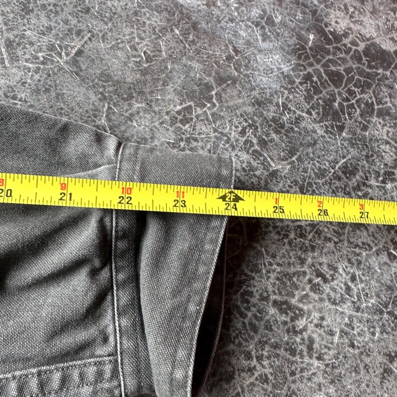 May include: Close-up of a dark gray denim jacket with a yellow measuring tape showing a length of approximately 24 inches. The jacket's fabric is visible, along with the seams and stitching.
