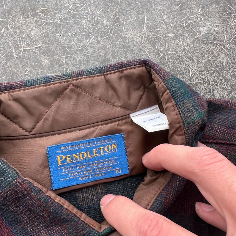 May include: Close-up of a Pendleton wool shirt with a plaid pattern in shades of burgundy, green, and brown. The shirt features a blue label with gold text that reads "Pendleton 100% Pure Virgin Wool Portland Oregon Since 1863".