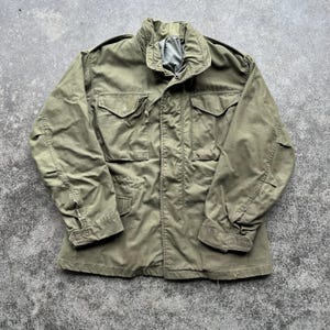 May include: An olive green military-style jacket with a high collar and multiple front pockets. The jacket has a button-up front and a relaxed fit. The fabric appears worn, suggesting a vintage or used condition. The jacket is laid flat on a textured surface.