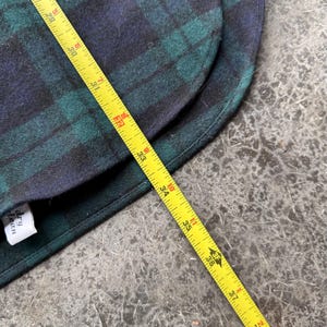 May include: A section of a green and navy blue plaid fabric, with a yellow measuring tape laid across it. The tape shows measurements up to 37 inches. A white tag with black text is visible.