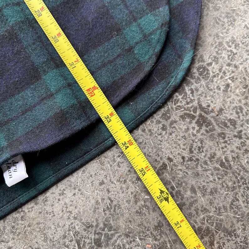 May include: A section of a green and navy blue plaid fabric, with a yellow measuring tape laid across it. The tape shows measurements up to 37 inches. A white tag with black text is visible.