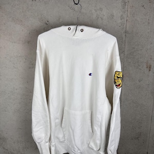 May include: A white long-sleeve hooded sweatshirt featuring a small embroidered Champion logo on the chest and a yellow tiger head patch on the left sleeve. The hoodie includes a front kangaroo pocket and two metal eyelets at the neckline.