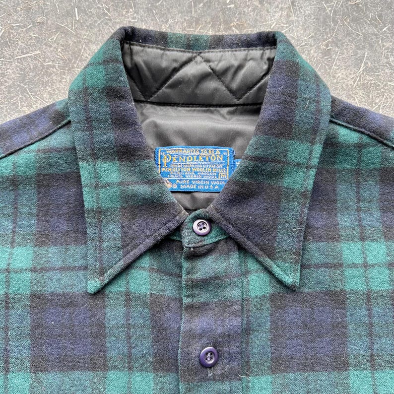 May include: A close-up of a green and navy plaid shirt with a black quilted collar. The shirt has a button-down front and a label that reads "Pendleton". The shirt is made of wool.
