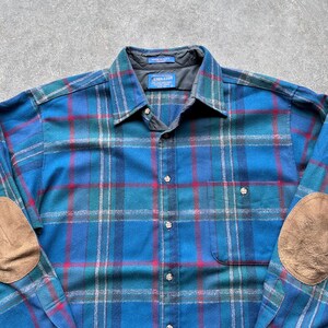 Op de afbeelding: Een blauw geruit overhemd met rode en groene accenten. Het overhemd heeft bruine elleboogstukken en een borstzak. De kraag heeft een klein label met "PENDLETON".