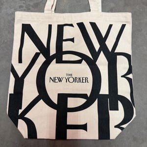 May include: A beige tote bag with black lettering that spells out "NEW YORKER". The words "THE NEW YORKER" are printed in a circle in the center of the bag. The bag has two beige handles.