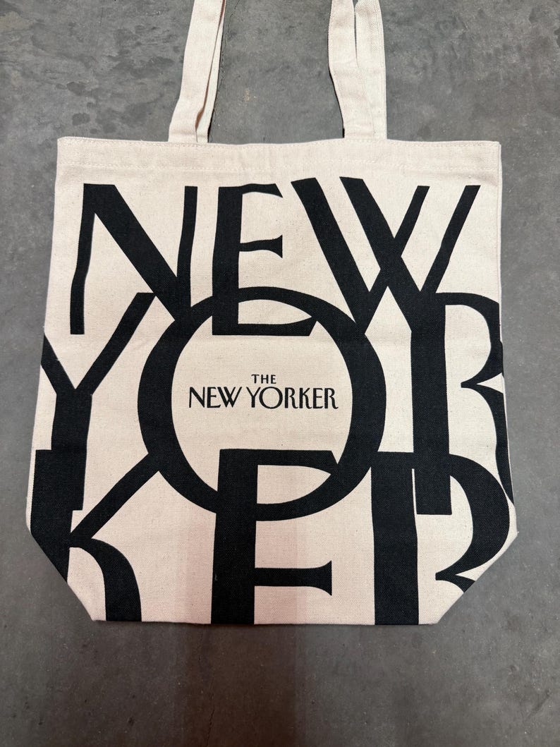 May include: A beige tote bag with black lettering that spells out "NEW YORKER". The words "THE NEW YORKER" are printed in a circle in the center of the bag. The bag has two beige handles.