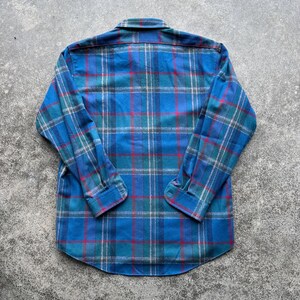 Op de afbeelding: Een blauw geruit overhemd met lange mouwen. Het ruitpatroon heeft tinten blauw, groen, rood en wit. Het overhemd heeft een klassieke kraag en manchetten met knopen. De stof lijkt een zachte flanel te zijn.