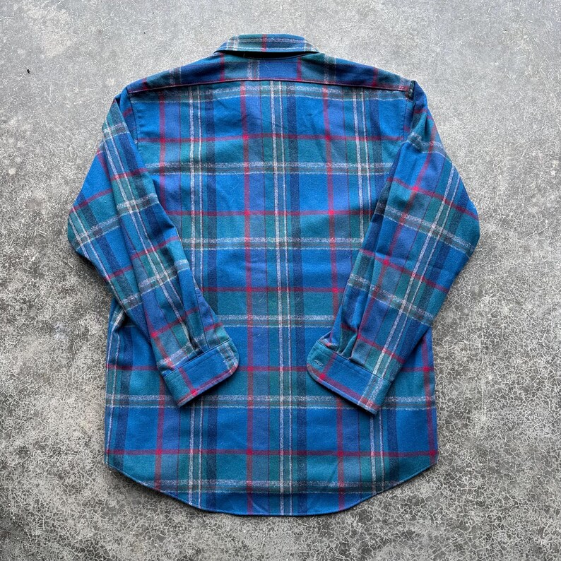 Op de afbeelding: Een blauw geruit overhemd met lange mouwen. Het ruitpatroon heeft tinten blauw, groen, rood en wit. Het overhemd heeft een klassieke kraag en manchetten met knopen. De stof lijkt een zachte flanel te zijn.