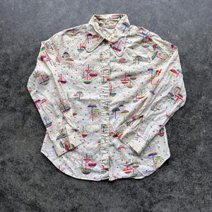 May include: Cream-colored button-down shirt with a colorful sailboat pattern. The long-sleeved shirt features a pointed collar and buttoned cuffs. The sailboats are in shades of red, blue, yellow, and green, with colorful polka dots.