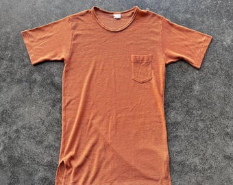 May include: An orange short-sleeved shirt with a chest pocket. The shirt has a ribbed texture and a crew neck. The shirt is a solid color and has a split hem on the sides.