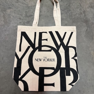 May include: A beige canvas tote bag with black lettering that spells out "NEW YORKER". The words "THE NEW YORKER" are in a black circle. The bag has two beige handles.