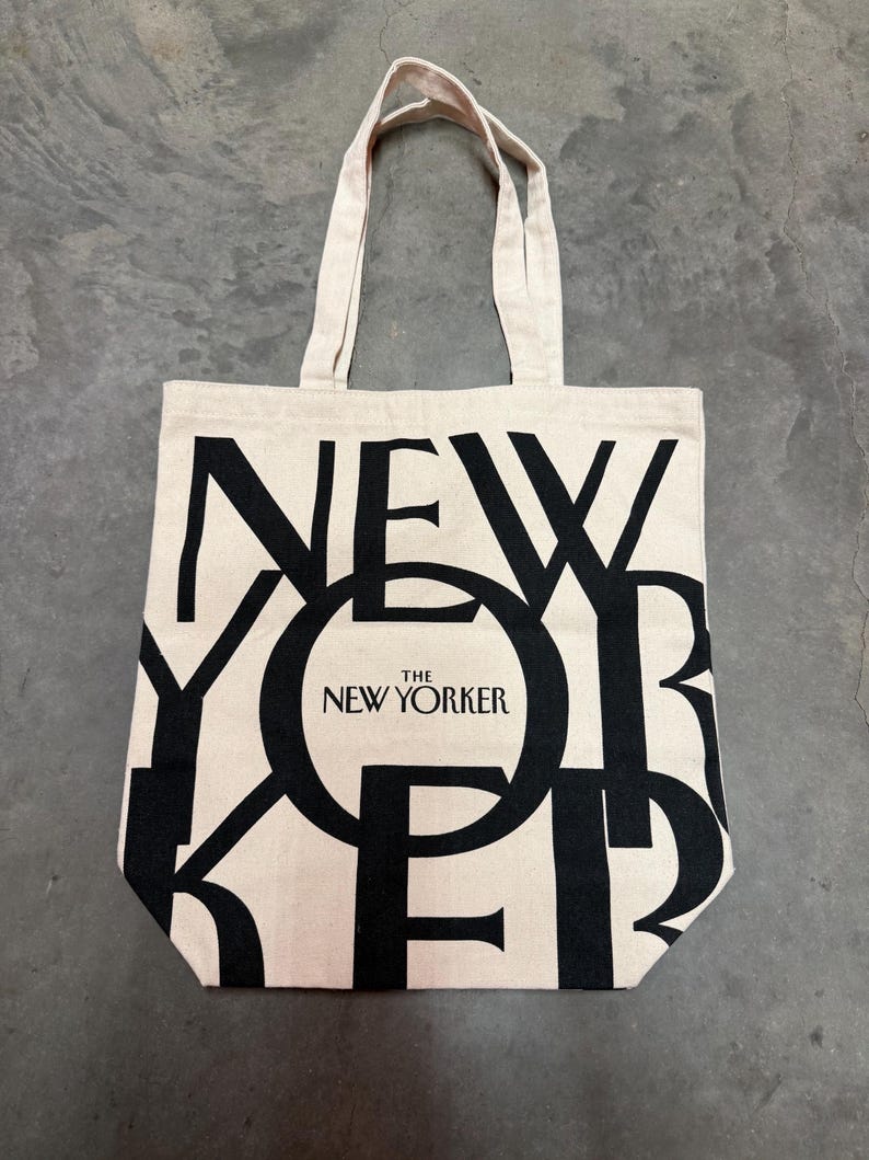 May include: A beige canvas tote bag with black lettering that spells out "NEW YORKER". The words "THE NEW YORKER" are in a black circle. The bag has two beige handles.