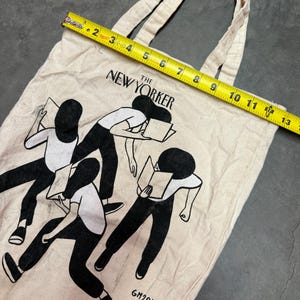 May include: Off-white canvas tote bag featuring a black and white illustration of figures reading books. The text "The New Yorker" is printed across the top. The bag measures approximately 13 inches in length.