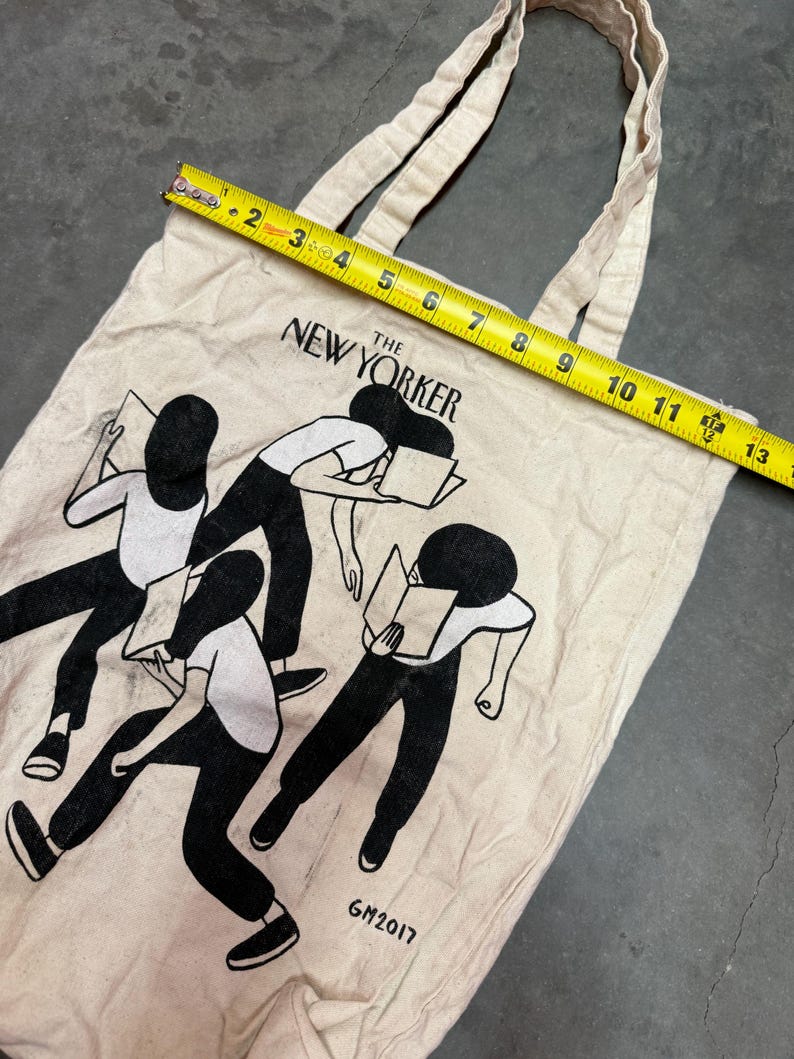 May include: Off-white canvas tote bag featuring a black and white illustration of figures reading books. The text "The New Yorker" is printed across the top. The bag measures approximately 13 inches in length.