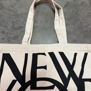 May include: Off-white canvas tote bag with black text that reads "NEW YORKER". The bag has two beige handles and is made of a sturdy fabric. The text is large and bold, making it easily readable.