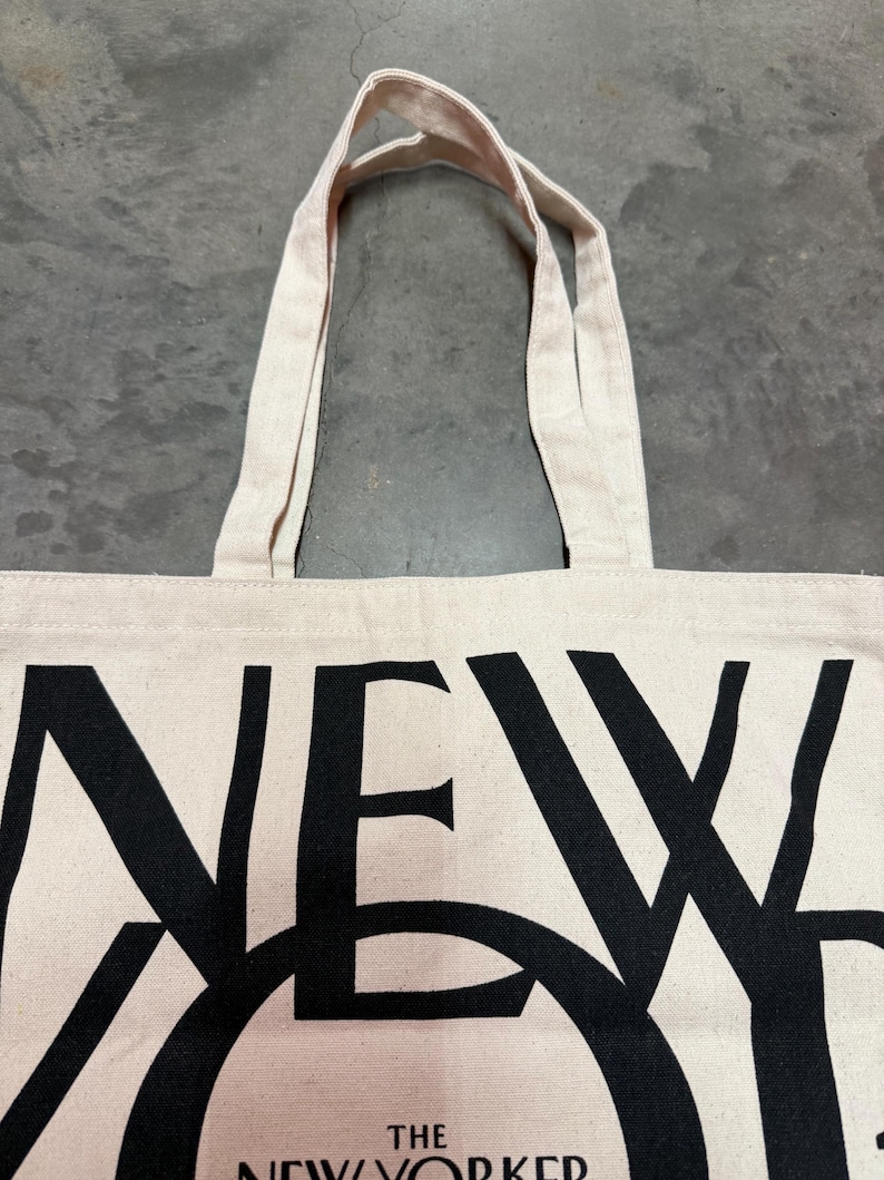 May include: Off-white canvas tote bag with black text that reads "NEW YORKER". The bag has two beige handles and is made of a sturdy fabric. The text is large and bold, making it easily readable.