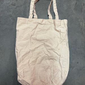 May include: A beige canvas tote bag with two matching handles. The bag is rectangular with a rounded bottom and shows signs of wear. The handles are attached to the top of the bag, allowing it to be carried over the shoulder or by hand.