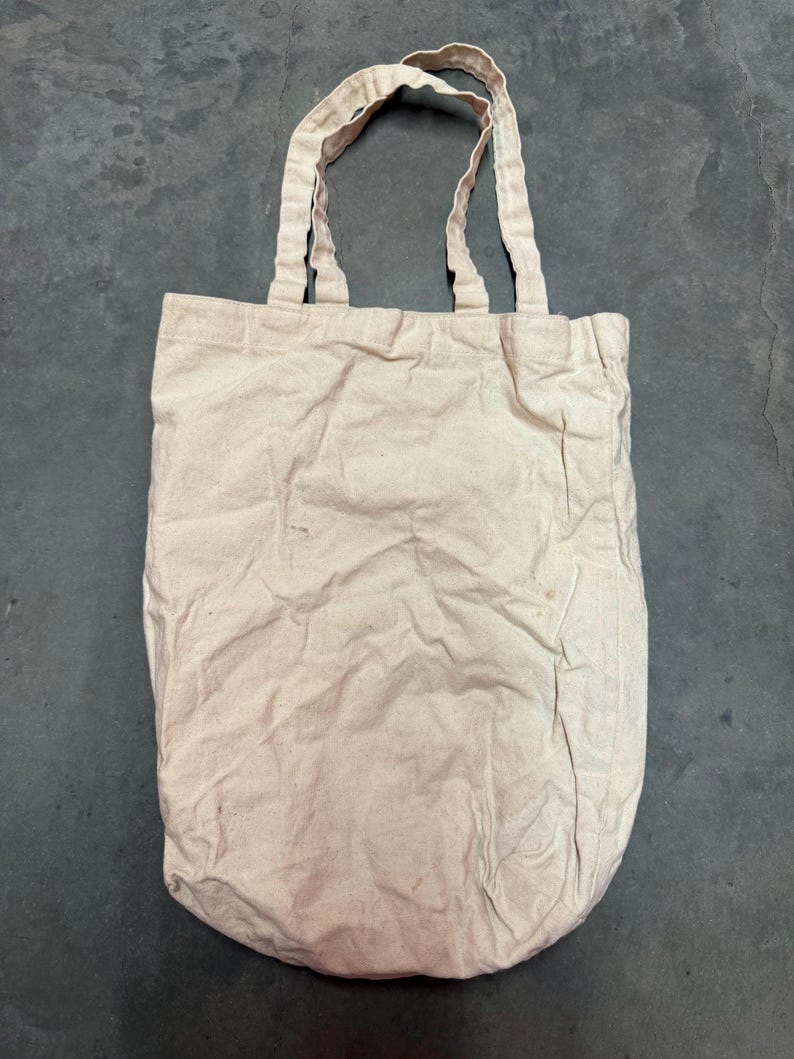 May include: A beige canvas tote bag with two matching handles. The bag is rectangular with a rounded bottom and shows signs of wear. The handles are attached to the top of the bag, allowing it to be carried over the shoulder or by hand.