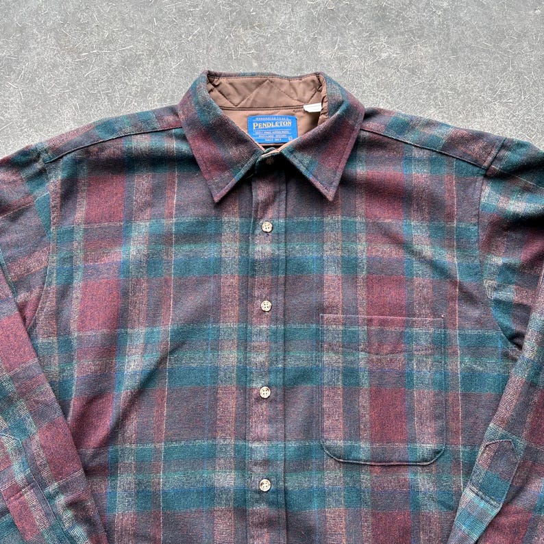 May include: A button-down plaid shirt in shades of teal, burgundy, and brown. The shirt features a collared neckline, a chest pocket, and the brand name "PENDLETON" on a tag. The fabric has a textured appearance.