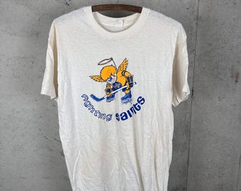 May include: A cream-colored short-sleeve t-shirt featuring a graphic of a cartoon angel with yellow hair and wings, wearing a blue and yellow hockey uniform while holding a hockey stick. The text "Fighting Saints" is printed in blue below the graphic.