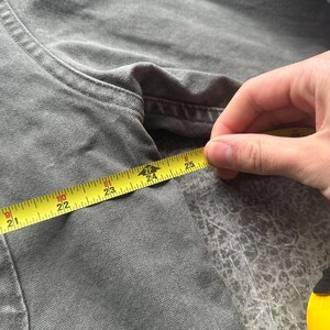 May include: Close-up of a gray garment being measured with a yellow tape measure. The tape measure shows a measurement of approximately 24 inches. The garment appears to be made of a textured fabric.