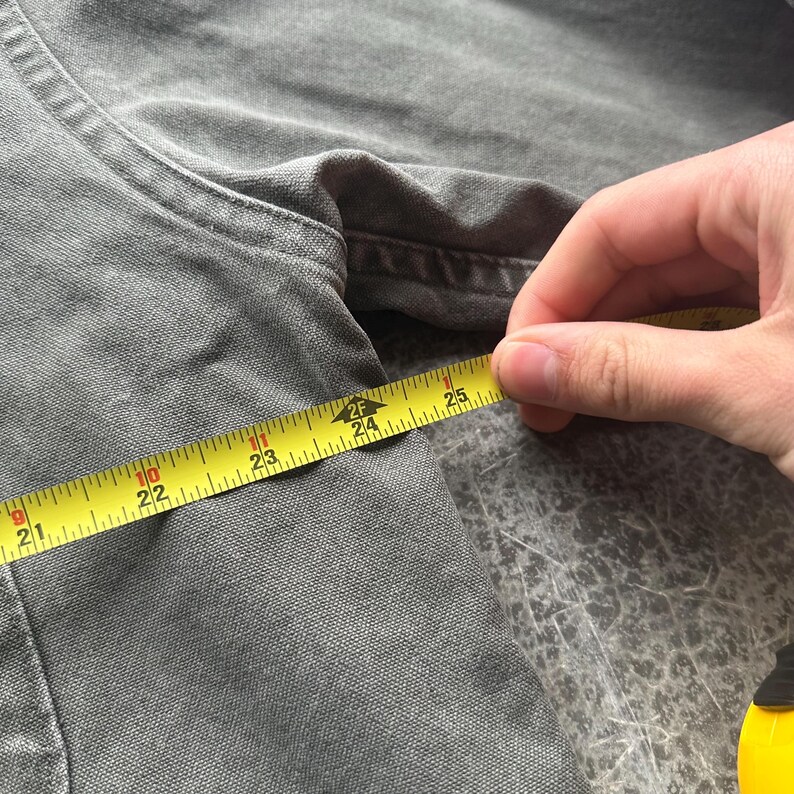 May include: Close-up of a gray garment being measured with a yellow tape measure. The tape measure shows a measurement of approximately 24 inches. The garment appears to be made of a textured fabric.