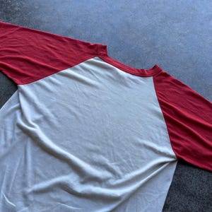 May include: A baseball style raglan shirt with a white body and red sleeves. The long-sleeved shirt is made of a soft material and is laid flat on a gray surface.
