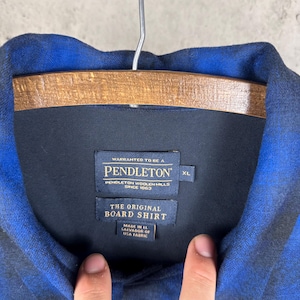 May include: A blue and black plaid long-sleeve shirt with a collar. The interior neck label reads: WARRANTED TO BE A PENDLETON, PENDLETON WOOLEN MILLS SINCE 1863, THE ORIGINAL BOARD SHIRT, MADE IN EL SALVADOR OF USA FABRIC, and the size tag indicates XL.
