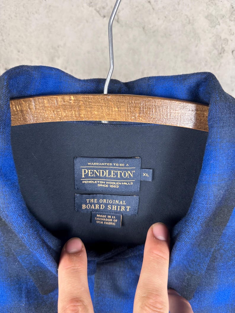 May include: A blue and black plaid long-sleeve shirt with a collar. The interior neck label reads: WARRANTED TO BE A PENDLETON, PENDLETON WOOLEN MILLS SINCE 1863, THE ORIGINAL BOARD SHIRT, MADE IN EL SALVADOR OF USA FABRIC, and the size tag indicates XL.
