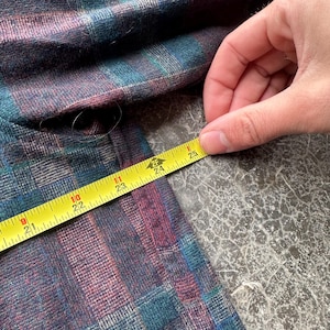 May include: A close-up of a plaid fabric in shades of teal, purple, and blue. A yellow measuring tape shows a length of approximately 24 inches. The fabric is on a gray surface.