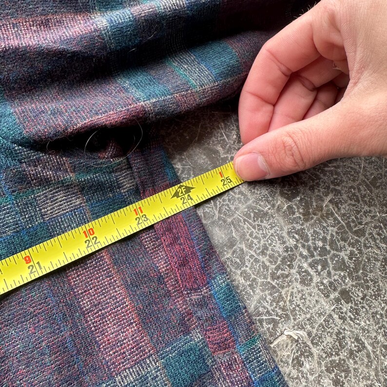 May include: A close-up of a plaid fabric in shades of teal, purple, and blue. A yellow measuring tape shows a length of approximately 24 inches. The fabric is on a gray surface.